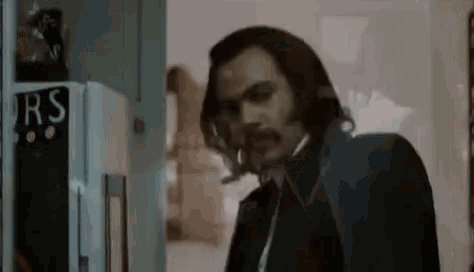 a man with long hair and a mustache is standing in front of a refrigerator in a dark room .
